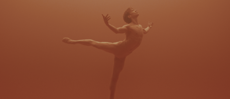 Daniil Simkin dances through a slightly orange, hazy atmosphere, posing in arabesque with his right leg lifted and his arm out to the sides, palms up. He arches his back and looks up towards the ceiling. He is bare chested and wears nude-colored tights and ballet slippers.