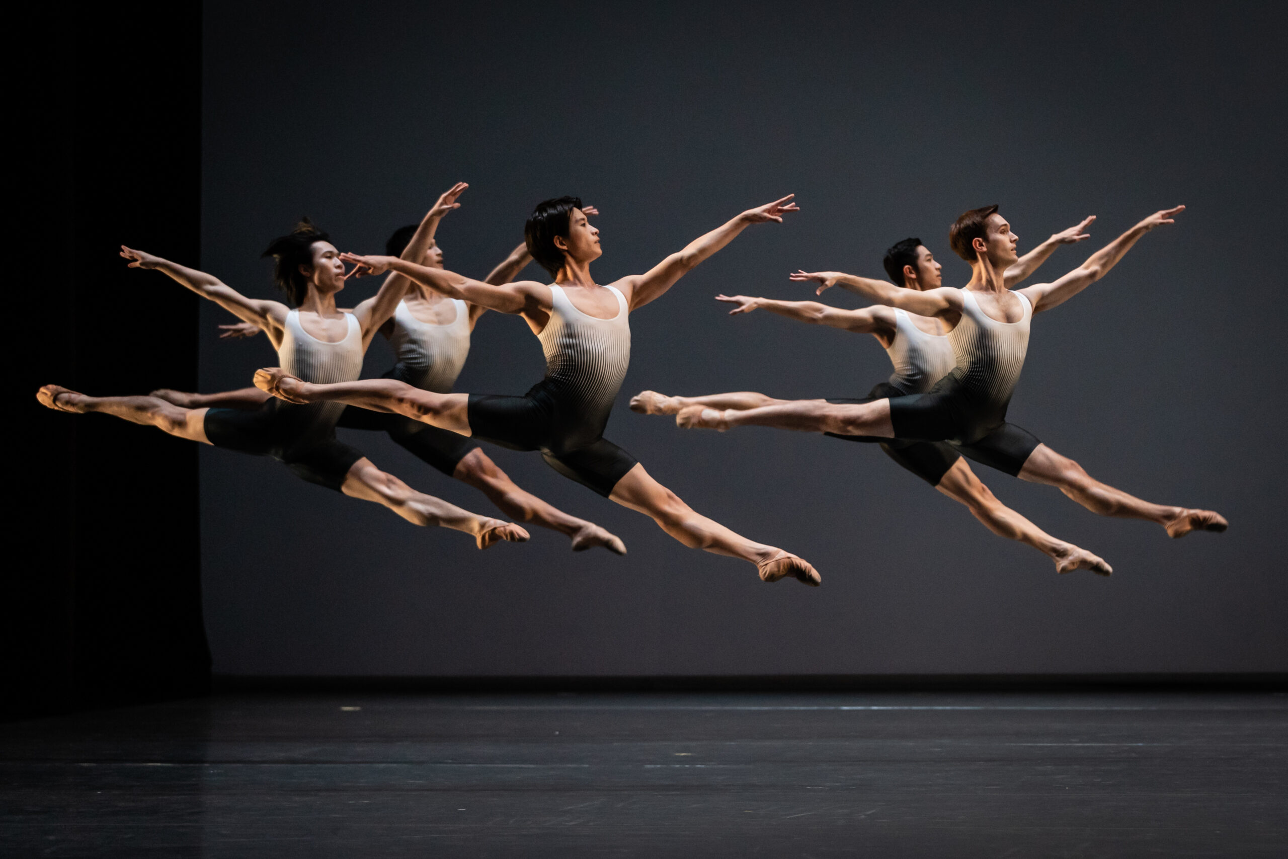 Six male members of Singapore Ballet are suspended mid-air in a split sissonne, wearing ombre white-to-grey biketards.