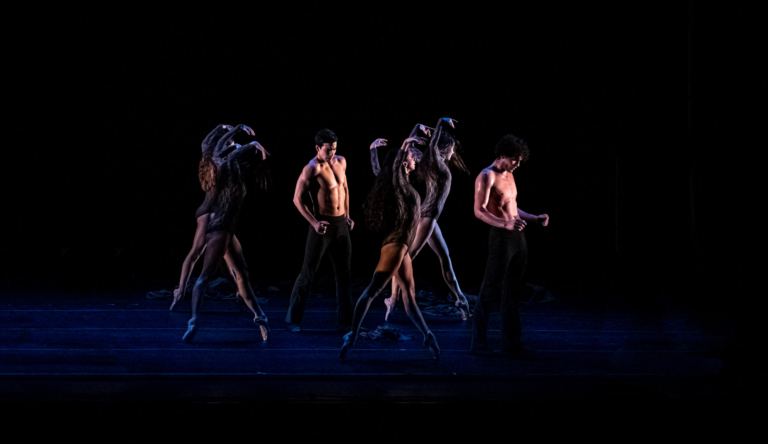 On a darkly lit stage, four female and two male dancers walk toward the right, the females on pointe and with their arms curving overhead.