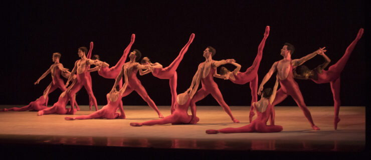 Fifteen members of The Washington Ballet, a mix of male and female dancers, pose together in one long block of bodies; some dancers stand in penché, others in a lunge tendu derriere, and others sitting with their left leg extended behind. They all wear red unitards.