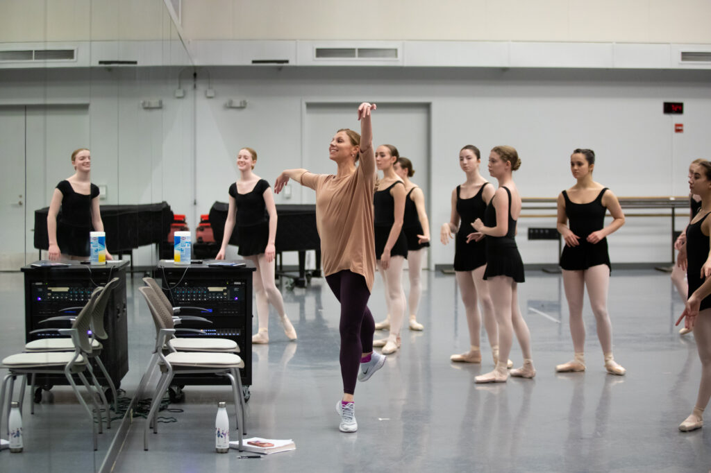 Lauren Fadeley Vayette, wearing an oversized mauve shirt, black leggings and sneakers, demonstrates a low first arabesque on demi-pointe. She looks sideways into the mirror. A large group of teenage female ballet students wearing black leotards, black ballet skirts, pink tights and pink pointe shoes, stand behind her, watching and smiling.