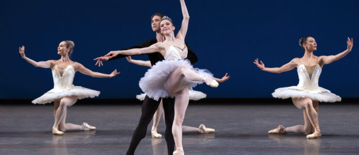 Megan Fairchild, in a white tutu and tiara, and Joseph Gordon, in black tights and tunic, perform a ballet pas de deux onstage. Three women in white tutus are behind them kneeling with their arms open. Fairchild does an attitude devant on pointe and rests her right hand on Gordon's extended right arm as he stands behind her in tendu, holding her around the waist with his left arm.