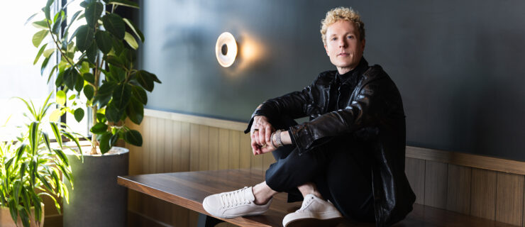 Remi Wörtmeyer sits cross-legged on a wooden table. He wraps his arms around his knees and clasps his hands. He wears a black leather jacket, black pants and white sneakers, and he looks toward the camera with a small smile.