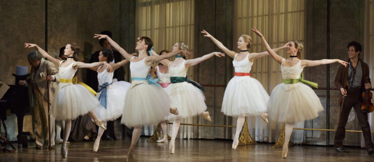 Six ballerinas in knee-length white tutus and brightly colored sashes do a temps levé arabesque towards stage right. They perform onstage in front of a dance studio set that includes two floor-to-ceiling arched windows covered in filmy drapes.