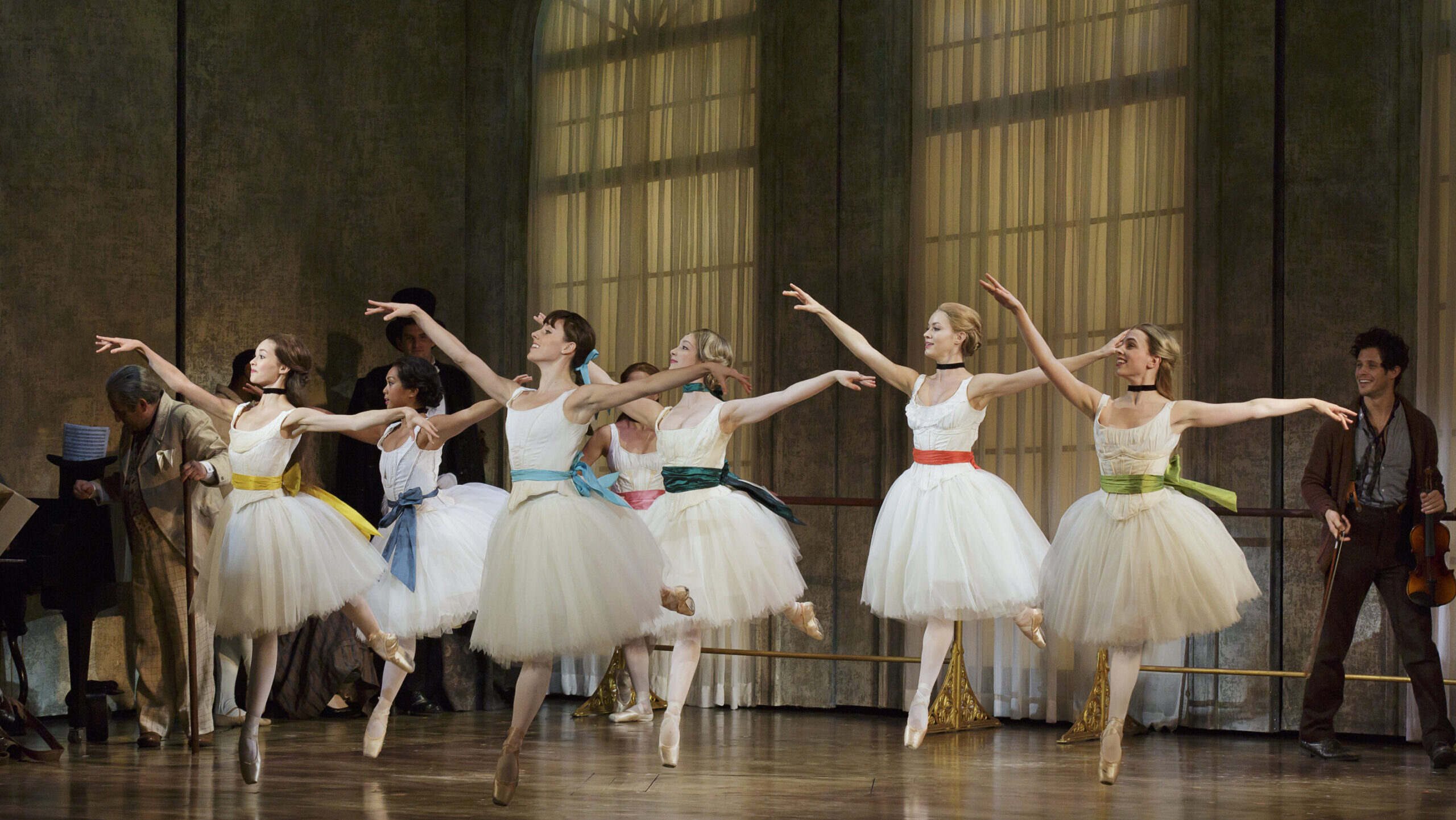 Six ballerinas in knee-length white tutus and brightly colored sashes do a temps levé arabesque towards stage right. They perform onstage in front of a dance studio set that includes two floor-to-ceiling arched windows covered in filmy drapes.