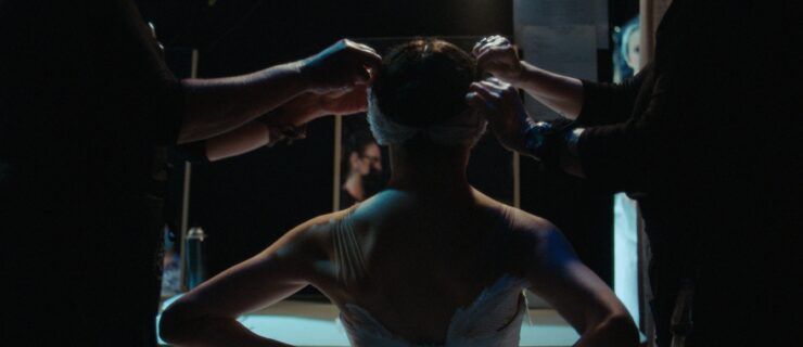 A female ballet dancer is shown backstage from the back. She sits down and hooks up her tutu bodice as two wardrobe assistants pin a feathered headpiece into her hair.