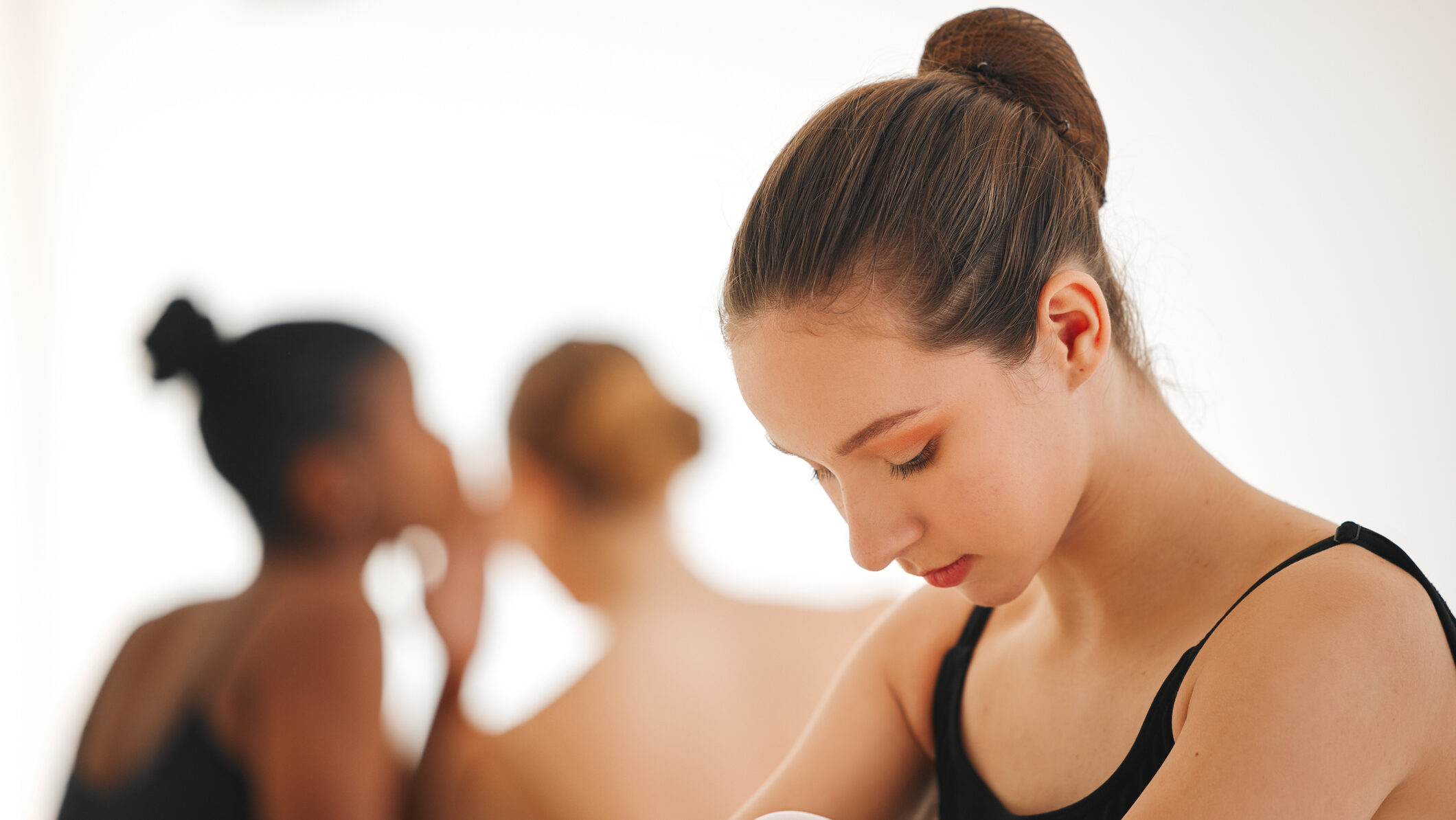 A teenage female ballet student wearing a black leotard and pink tights is shown from the chest up, sitting on the ground with her knees tucked up and her arms wrapped around them. She looks down at the floor as two other teenage female dancers, blurred and sitting in the background, gossip and whisper to each other.