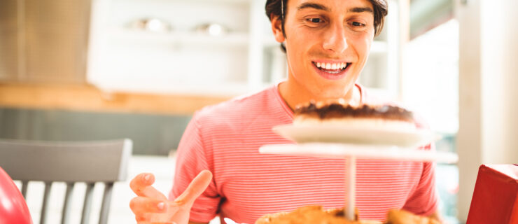 A young man in a pink long-sleeved shirt smiles as he reaches toward a platter of cookies.