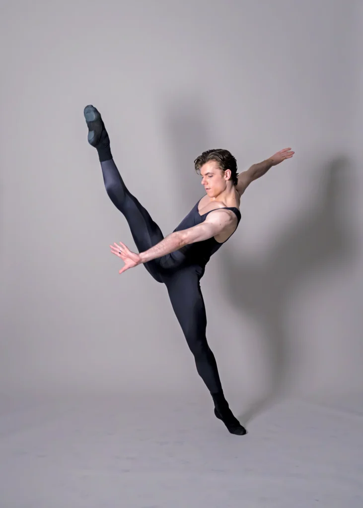 Jake Roxander, wearing a black unitard, does a high grand battement devant with his right leg. He reaches his left arm down and in front of him, hand flexed, and his right arm behind him. He dances in front of a gray backdrop and looks down towards his left hand.