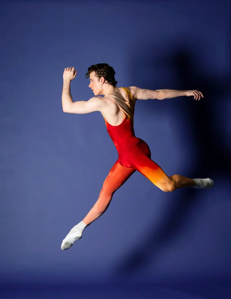 Jake Roxander, wearing a unitard in red and orange hues, jumps up in front of a blue backdrop. He faces stage right, and his right leg is straight and his left leg is bent in attitude derrierre. His lleft arm is bent and frames his face, while his right arm is stretched back behind him.