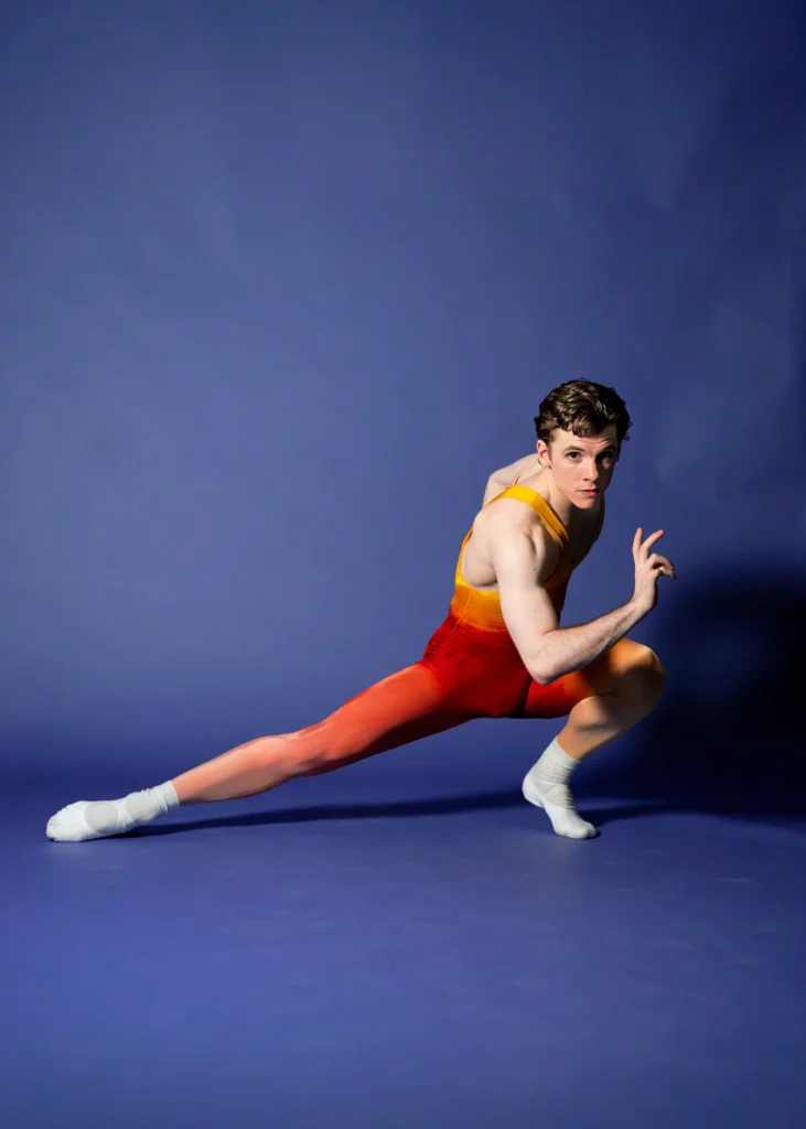 Jake Roxander crouches low to the ground and stretches his right leg out to his side. He wears a unitard in red and orange hues, white socks and white ballet slippers. He bends his right arm in front of him with splayed fingers and looks intensely at the camera. He poses in front of a blue backdrop.