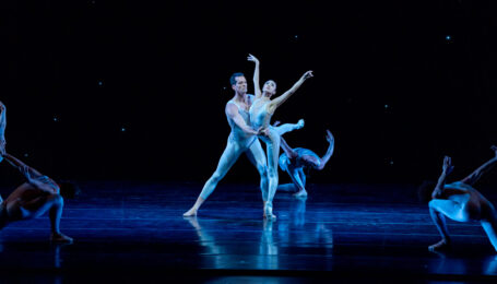 Joffrey Ballet's Jeraldine Mendoza and Dylan Gutierrez perform a pas de deux in white unitards, surrounded by four dancers kneeling and leaning toward them. The stage is dark, lit with a blue light, with a starry night sky effect in the background. Mendoza stands in arabesque en pointe, arms lifted to the sky and eyes up, as Gutierrez supports her around the waist and stands in a side lunge, looking at her.