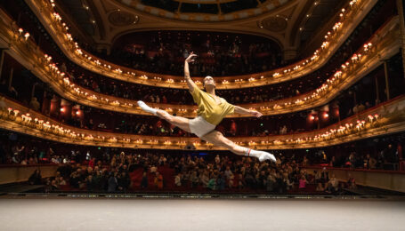 Joseph Sissens flies in a grand sissone onstage during company class, the theater house behind him. He wears a yellow shirt and white shorts.