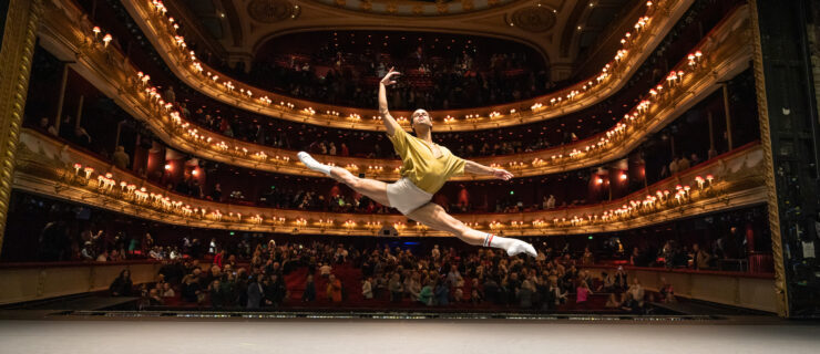 Joseph Sissens flies in a grand sissone onstage during company class, the theater house behind him. He wears a yellow shirt and white shorts.