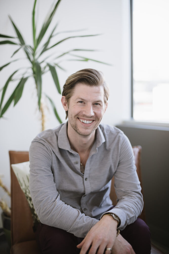 Josh Spell, a man approximately in his thirties, sits in a chair and looks straight at the camera, resting his forearms on his thighs and crossing his hands. He wears a gray button-down shirt and dark pants. He sits in front of a window and a small indoor tree.