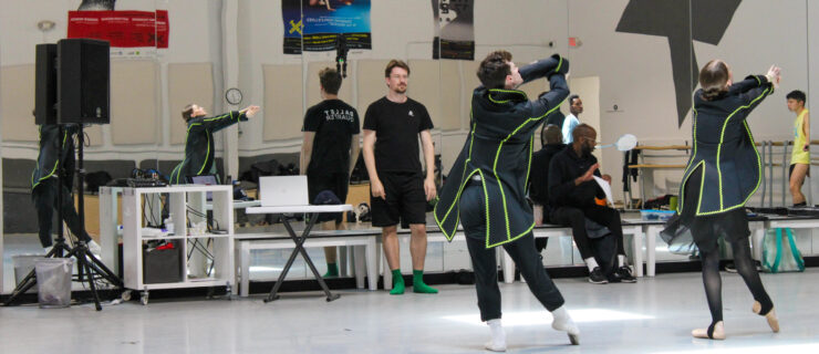 In a large studio with dance posters on the back wall, Loughlan Prior leads rehearsal as two dancers tendu together in long black waistcoats with neon green trim.