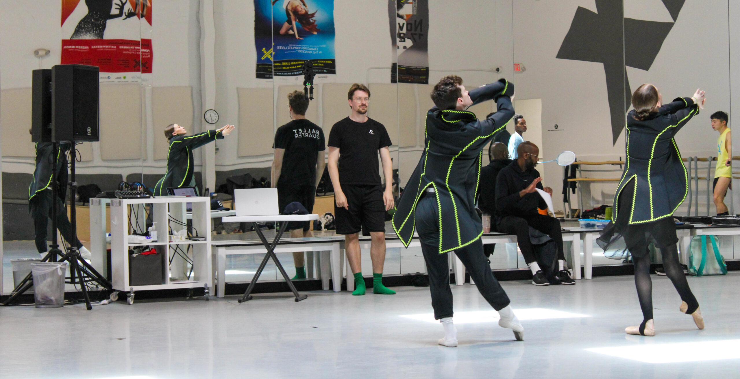 In a large studio with dance posters on the back wall, Loughlan Prior leads rehearsal as two dancers tendu together in long black waistcoats with neon green trim.