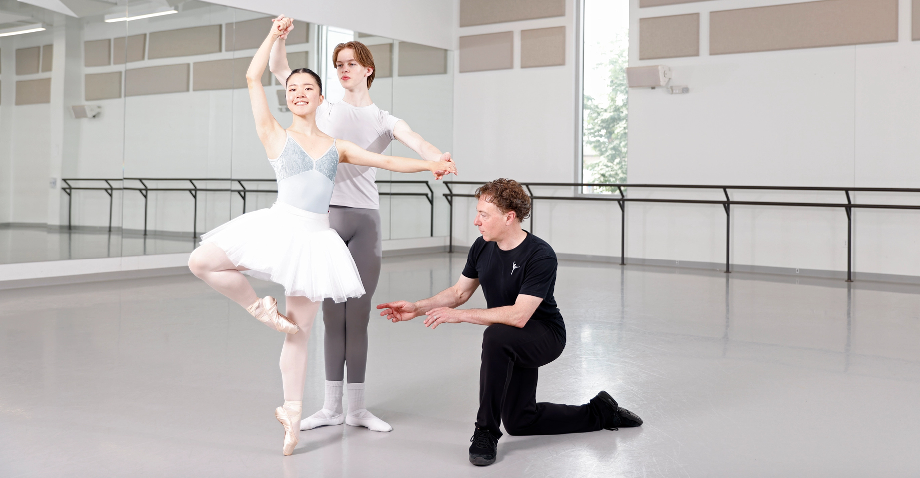 A male and female ballet student practice finger turns in a large dance studio. Their teacher, wearing a black t-shirt, workout pants, and sneakers, kneels on the ground next to them and points to the female dancer's retiré position on pointe. The girl wears a light-blue leotard, white practice tutu, pink tights and pointe shoes, while her partner wears a white t-shirt, gray tights, and white socks and ballet slippers.