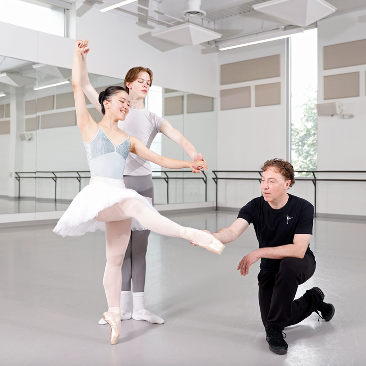 Two teenage ballet students, a boy and girl, practice finger turns in a large dance studio. The boy, in a white t-shirt and gray tights, holds the girl's hands as she does a développé devant, her knee not quite stretched yet. A male teacher, wearing a black t-shirt and workout pants, kneels next to them on their left and holds her pointed right foot.