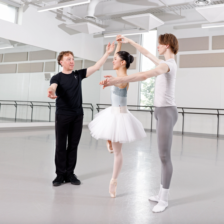 A male ballet teacher instructs a male and female ballet student (both in profile) on finger turns in a large dance studio. The girl, who wears a tutu, balances on pointe with her right leg in retiré and holds her arms in third position, while the boy stands behind her and holds her hands. The teacher stands in front of them and lifts his arms slightly up, smiling. He wears a black t-shirt and workout pants, while the students wear practice clothes.