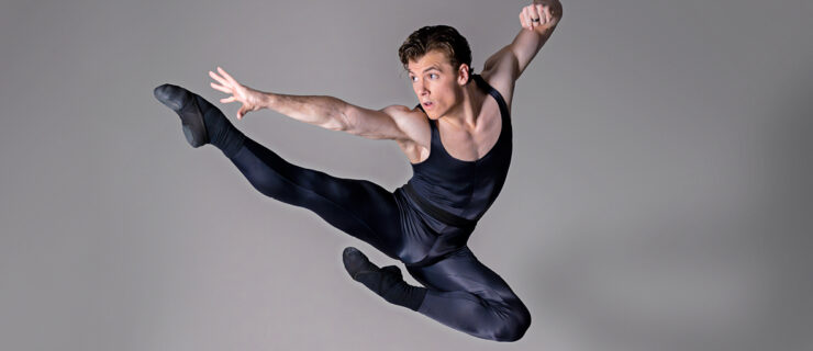 Jake Roxander, wearing a black unitard, does an Italian pas de chat in front of a gray backdrop. He pulls his left arm back and makes a fist, and stretches his right arm out.