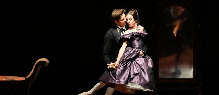 Fredmann Vogel lunges forward onto hi s left leg, holding Elisa Badenes around her waist as she does a small arabesque on pointe on her left leg. She pushes her hips forward and leans her upper body against Vogel's, turning her face towards him so that their cheeks touch. She wears a 19th-century-style purple off-the-shoulder dress with a full skirt, while Vogel wears a black smoking jacket, black tights and ballet slippers, white shirt and black tie. They dance onstage in front of a loveseat and mirror.