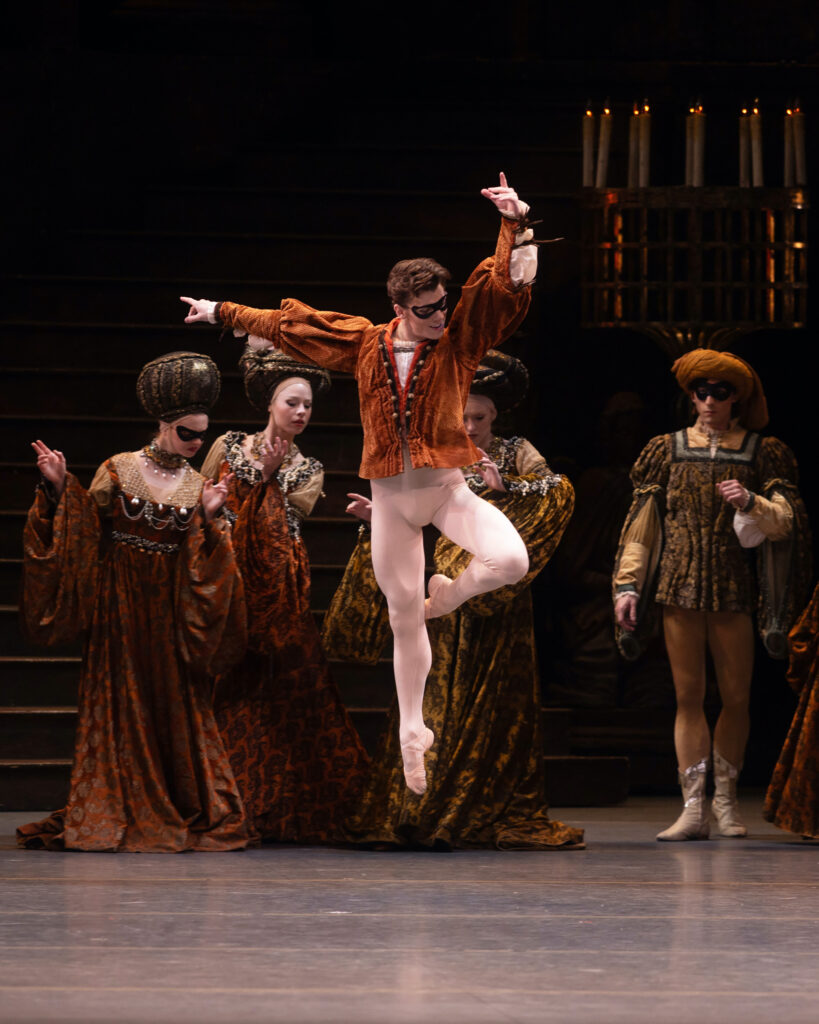 Jake Roxander jumps high into the air with his left leg in passé. His right arm is to the side and his left arm is up, and he looks down towards his left leg with a confident smile. He wears white tights and ballet slippers, a rust-colored tunic, and a black mask over his eyes. Behind him, dancers in Renaissance court costumes watch him.