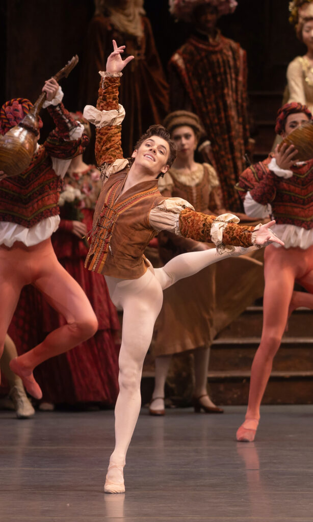 Jake Roxander performs onstage, taking an attitude derierre with his right leg lifted. He cocks his chin up slightly and smiles confidently. He wears white tights and ballet slippers and a rust-colored tunic; behind him, a crowd of dancers in Renaissance costumes mill about and dance.