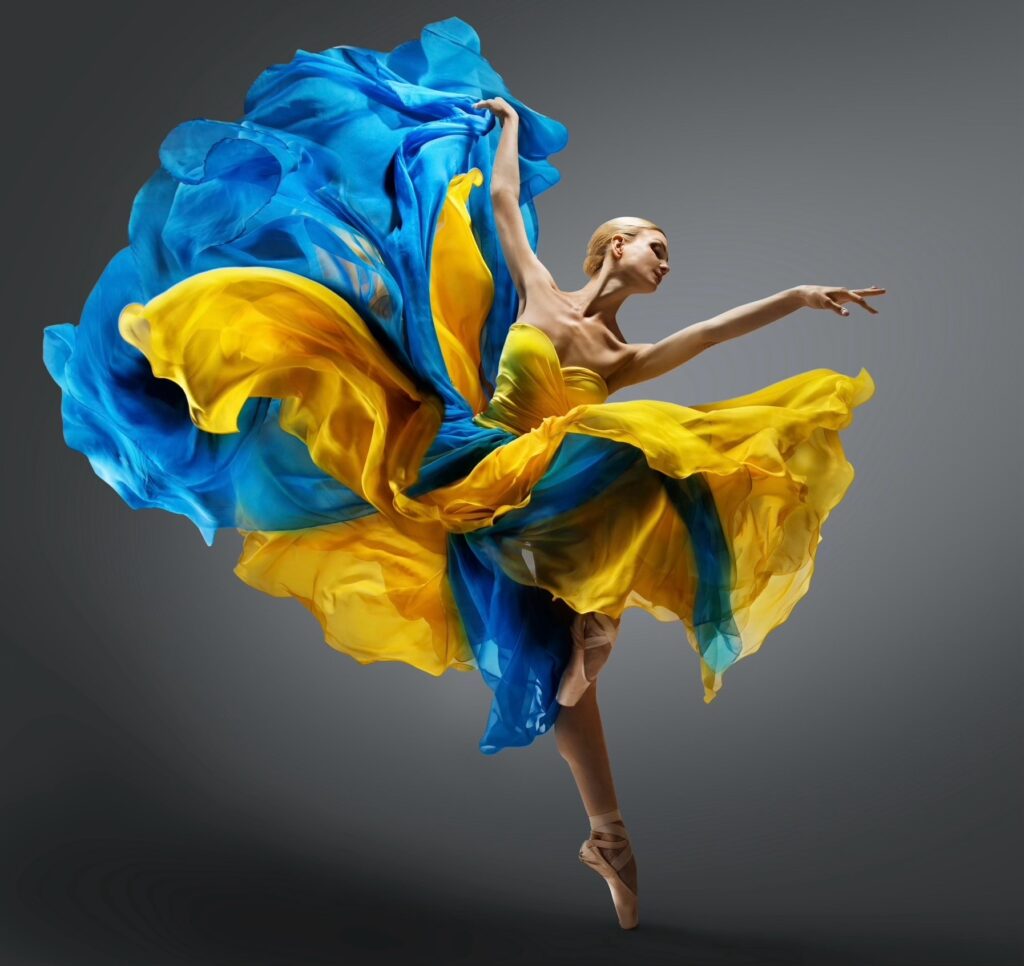 A blond ballerina relevés onto her left foot with her right leg in a parallel retiré position, lifting her right arm up and her left arm out in front of her. She wears a long, billowy blue and yellow dress and pointe shoes.