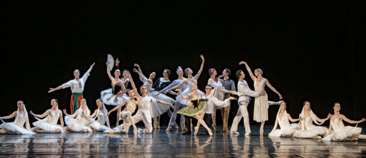 Members of the National Ballet of Ukraine pose in a tableau onstage wearing various costumes. The front line of dancers take poses on or low to the floor, while the back line pose standing or on pointe. The are in front of a black backdrop.