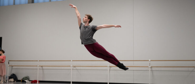 In a large ballet studio, Jack Thomas suspends in the air in a cabriole derriere, his arms in a high arabesque position. We wears a gray t-shirt and maroon tights.