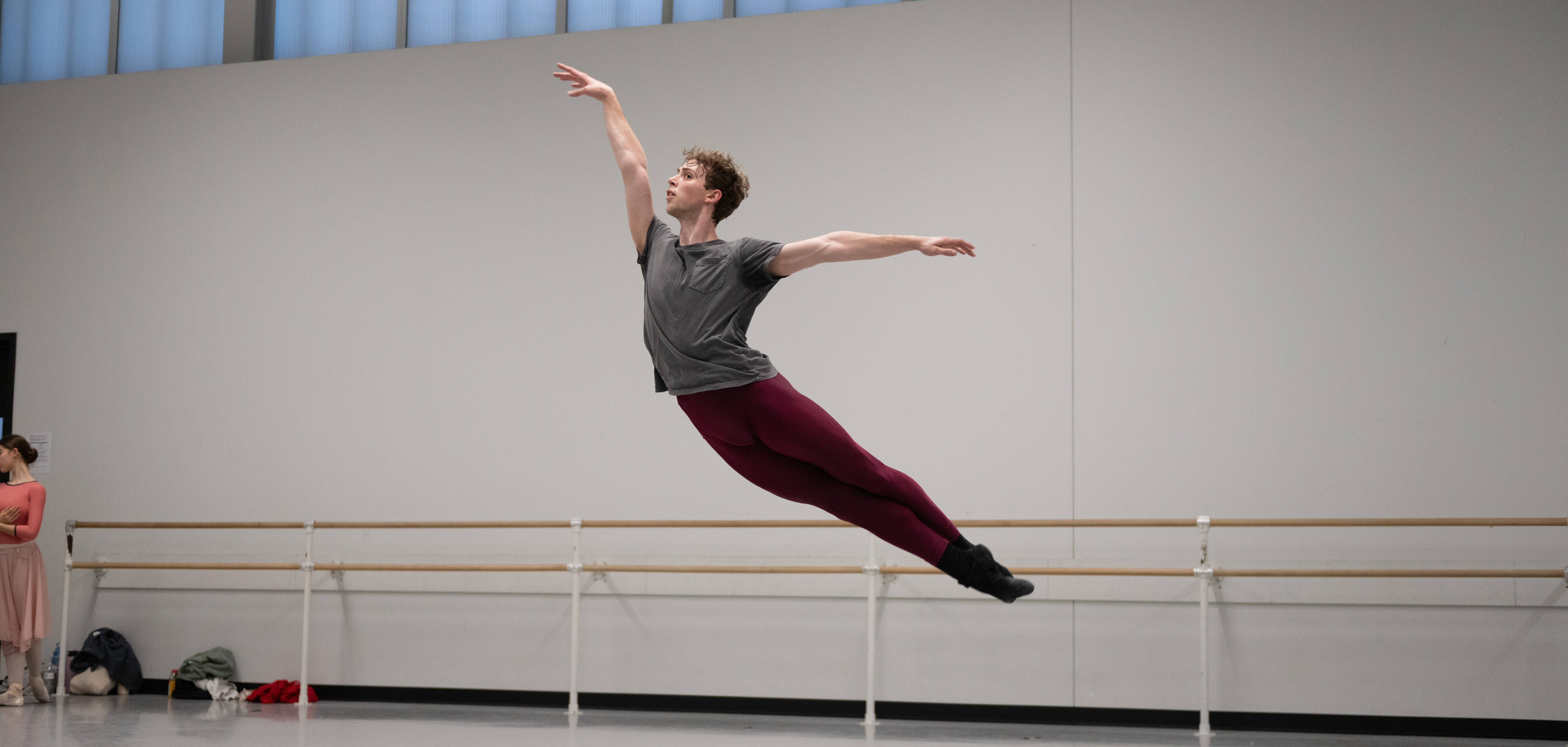 In a large ballet studio, Jack Thomas suspends in the air in a cabriole derriere, his arms in a high arabesque position. We wears a gray t-shirt and maroon tights.