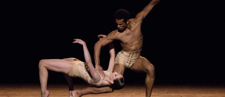 Jack Gasztowtt and Valentine Colasante dance a pas de deux from Jiří Kylián’s Petite Mort. They wear minimalist flesh-tone costumes. Colasante bridges back across Gasztowtt's back leg as he lunges forward, left arm extended and right gently holding Colasante's left shoulder.