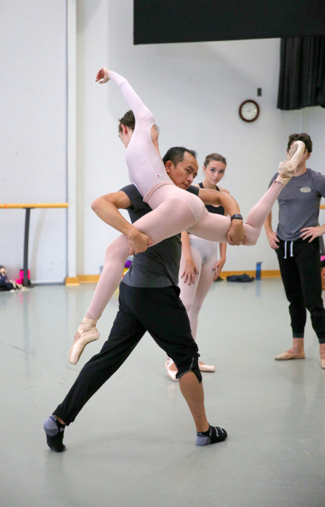 Ma Cong partners a female dancer as he leads rehearsal, holding her in a split attitude position as he supports her below the knees.