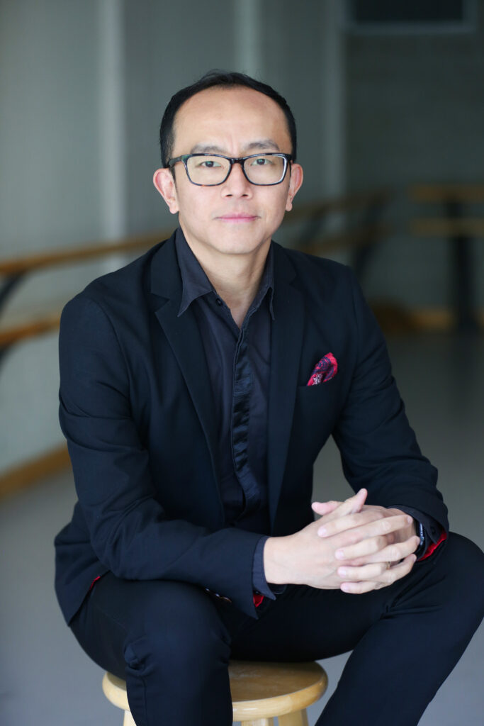 Ma Cong wears a black tuxedo and sits on a light wooden bench, posing for a headshot. He smiles slightly.