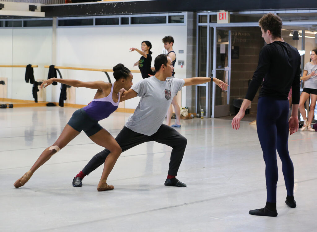 Ma Cong dances with a female Richmond Ballet company member in rehearsal as he leads rehearsal. They both lunge and lean to the left, a male dancer watching them closely.