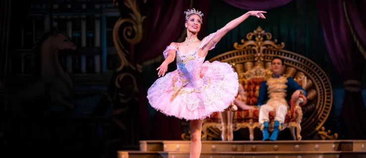 Katlyn Addison does a piqué attitude in croisé and sweeps her arms to her left, her left arm raised and her right slightly lower. She looks out over her left shoulder and smiles elegantly. She wears a pink tutu with gold and rhinestone embellishments, a jeweled necklace and tiara, brown tights and brown pointe shoes. Behind her, a young male and female dancer sit on a throne and watch her dance.