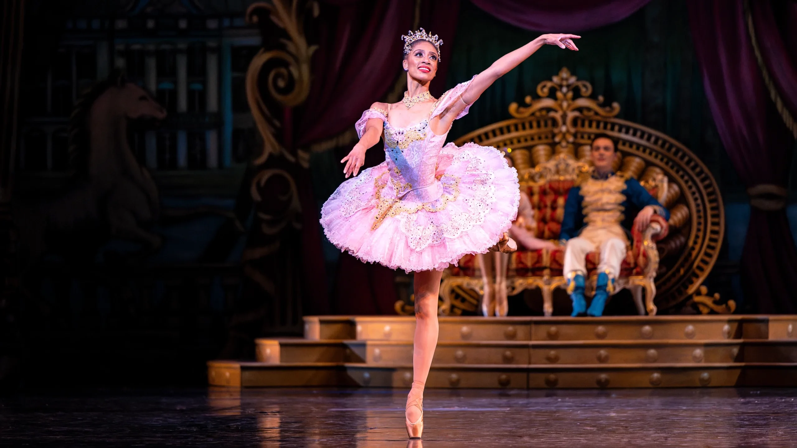 Katlyn Addison does a piqué attitude in croisé and sweeps her arms to her left, her left arm raised and her right slightly lower. She looks out over her left shoulder and smiles elegantly. She wears a pink tutu with gold and rhinestone embellishments, a jeweled necklace and tiara, brown tights and brown pointe shoes. Behind her, a young male and female dancer sit on a throne and watch her dance.