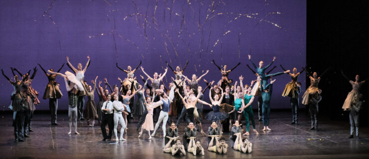 A large ensemble of ballet dancers pose together onstage in jubilant celebration. Gold streamers and confetti fly through the air in front of the deep purple backdrop. The dancers wear glitzy costumes, including pancake tutus.