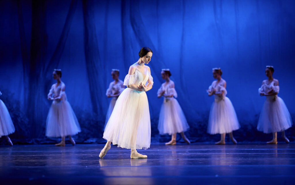 A ballerina in a long white tutu poses center stage. She is in profile facing stage left with her right leg in tendu derrierre and her wrists crossed in front of her chest, her upper body slightly pitched forward and her eyes down towrads the floor. In the background, a diagonal line facing the downstage right corner of women in the same costume and with white flower wreaths in their hair pose with their left foot in b plus effacé, their arms also crossed in front of their chest.