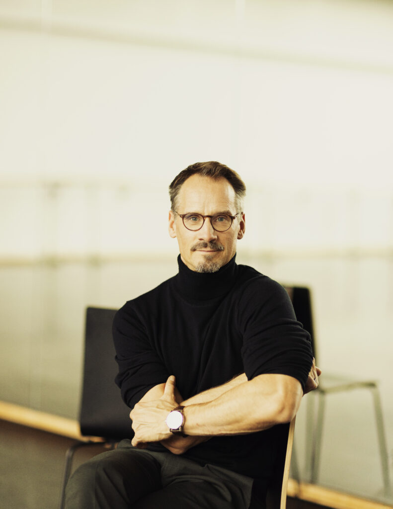 Christian Spuck, shown from the waist-up, sits on a chair in a ballet studio with his back to the mirror. He wears a black turtleneck and glasses, and he crosses his arms confidently as he smiles gently at the camera.