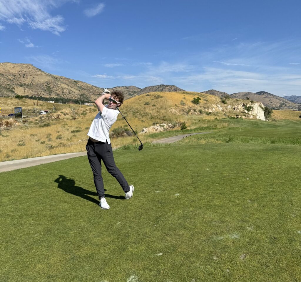 Jack Thomas, wearing a white t-shirt and black pants, prepares to swing his club on a golf course.