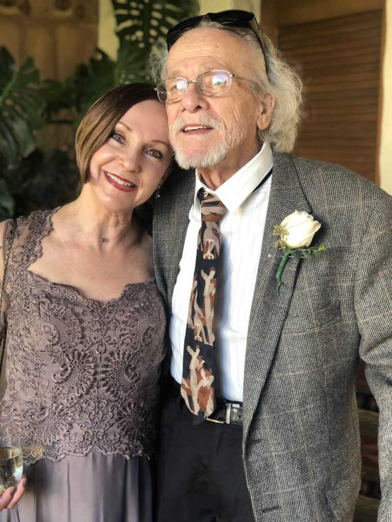 James Clouser, dressed in a tweed sport jacket, white shirt, black and brown tie and black pants, puts his right arm around Melissa Lowe, who is wearing a purple evening gown with a lace bodice. He looks to the right as she looks at the camera and smiles.