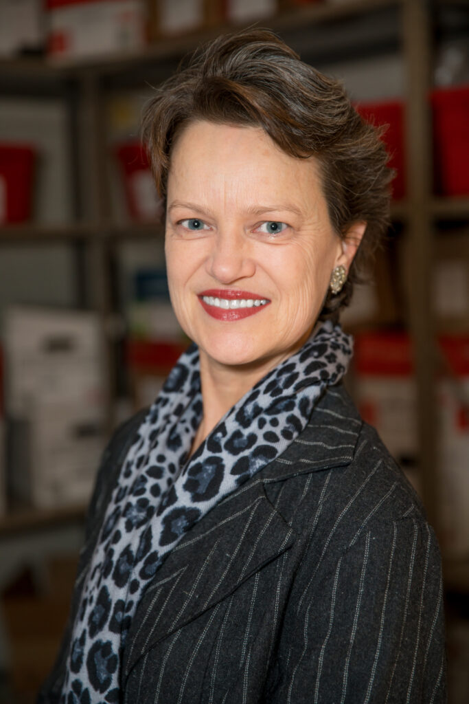 Kathleen Mitchell is shown from the chest up. She wears a pinstriped blazer with a blue leopard-print scarf. She looks directly towards teh camera and smiles.