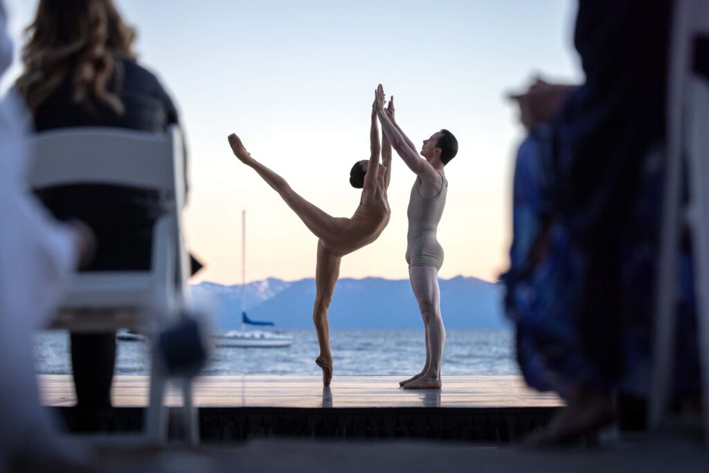 Lia Cirio does a high arabesque on pointe with her right leg lifted. Her partner, Paul Craig, stands in front of her. They both lift their arms up and press their hands together. Cirio wears a tan leotard and Craig wears a buff-colored biketard. They perform on an outdoor stage with Lake Tahoe in the background. A sailboat sails by.