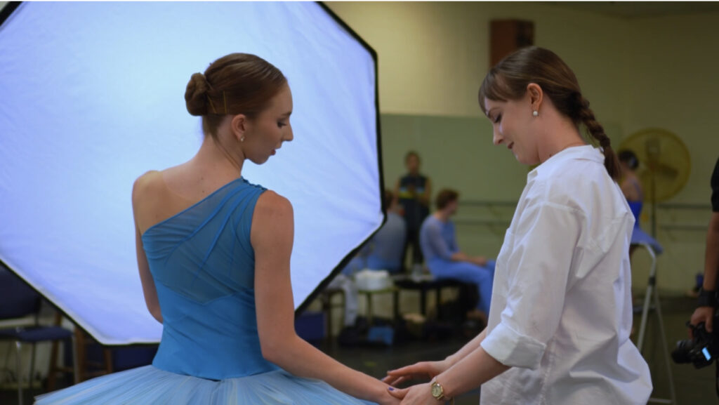 Margaret Mullin stands to the right of a ballerina in a blue tutu during a photo shoot. She lightly touches the dancers right hand and smiles. Mulling wears a white button-down blouse. They stand in a dance studio with a large photography umbrella in the background.