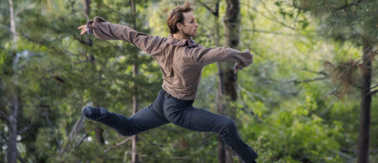 Adrian Danchig-Waring performs ballet on an outdoor stage wearing a brown long-sleeved shirt and blue pants and ballet slippers. He jumps up in profile facing stage left, with both legs in an elongated attitude position and his arms out. The stage is surrounded by pine trees.