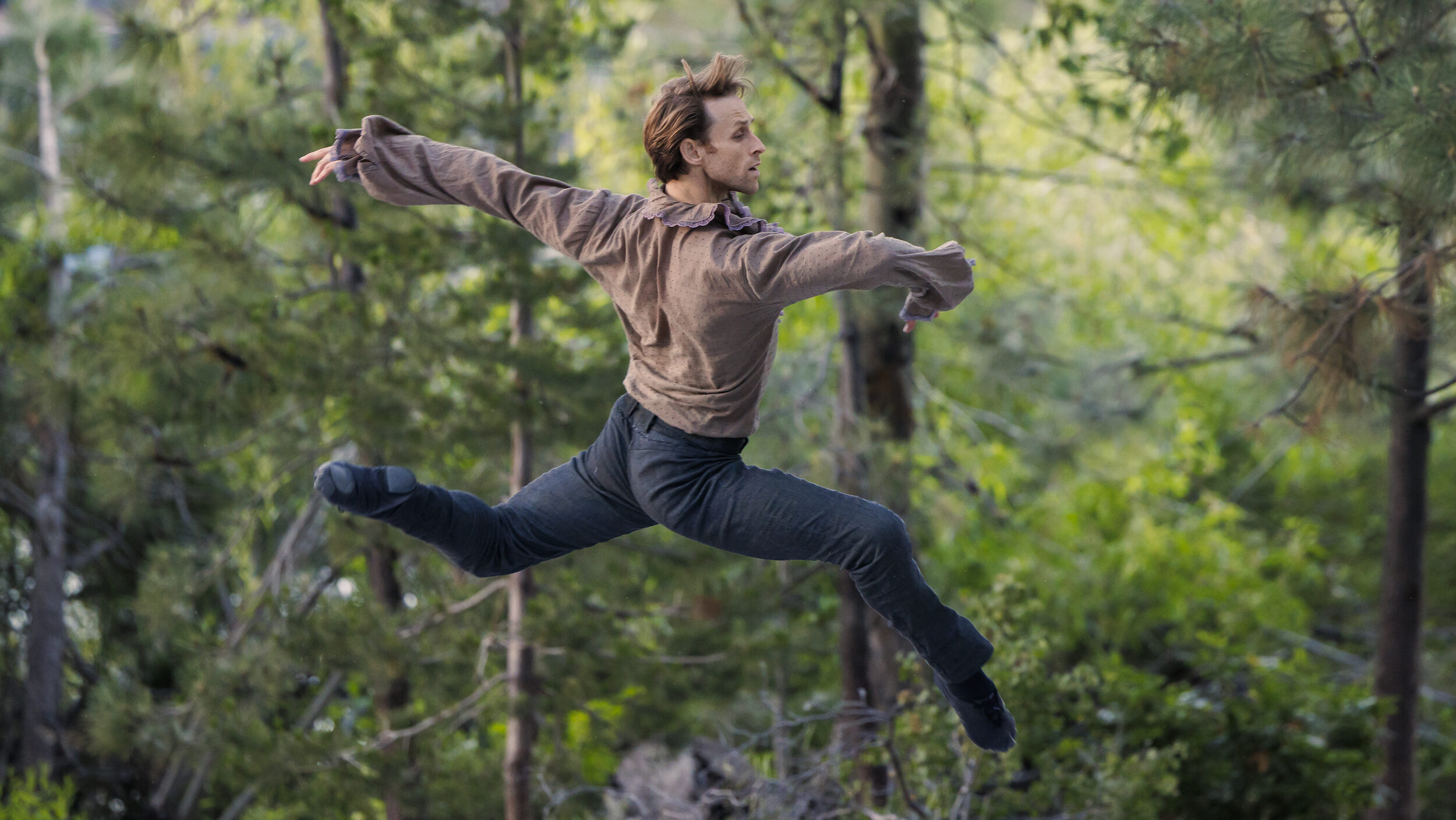 Adrian Danchig-Waring performs ballet on an outdoor stage wearing a brown long-sleeved shirt and blue pants and ballet slippers. He jumps up in profile facing stage left, with both legs in an elongated attitude position and his arms out. The stage is surrounded by pine trees.