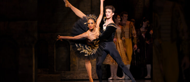 Katlyn Addison and David Huffmire perform a pas de deux onstage. Huffmire lunges to his right and holds Addison by the waist as she lunges forward in a penché in attitude, arms out to the side and pressed slightly behind her. She wears a black tutu, brown tights and pointe shoes, and a large, golden tiara, and she looks out towards the audience with a powerful demeanor. Huffmire is costumed in black tights and ballet slippers, and a black velvet tunic.