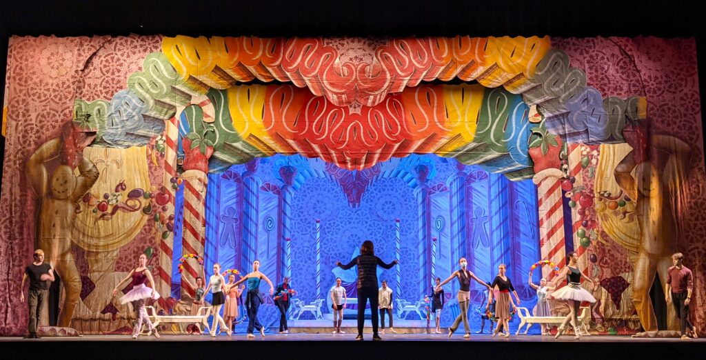 A company of ballet dancers stand onstage during a Nutcracker rehearsal under a set depicting the Land of Sweets. They wear various rehearsal clothes and stand in a triangular-shaped formation. Margaret Mullin, her back to the camera, stands center stage and gives the dancers notes.